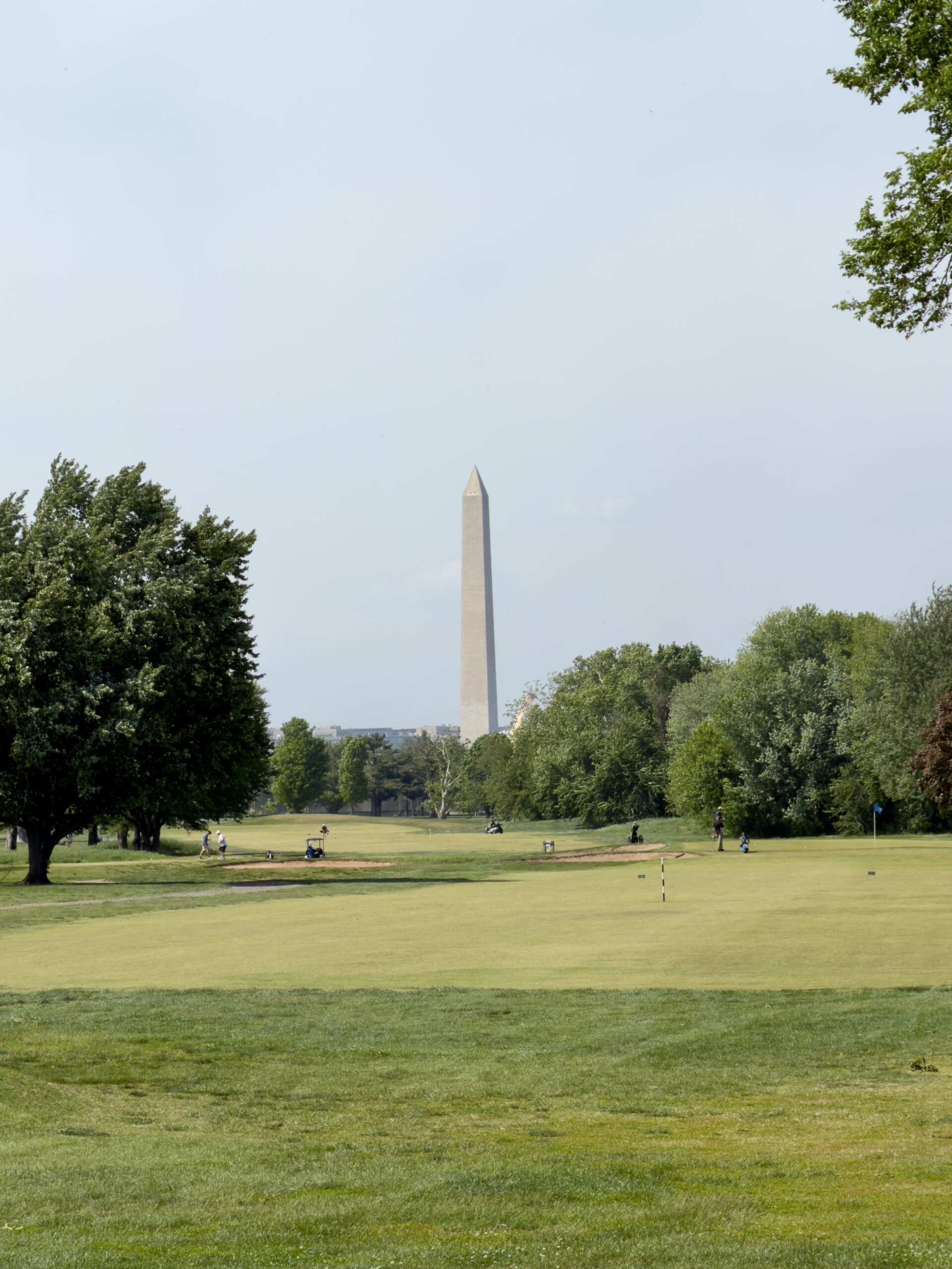 Washington Monument framed by trees beyond the 16th fairway at East Potomac Golf Links Blue Course, viewed from the tee box on a spring morning.