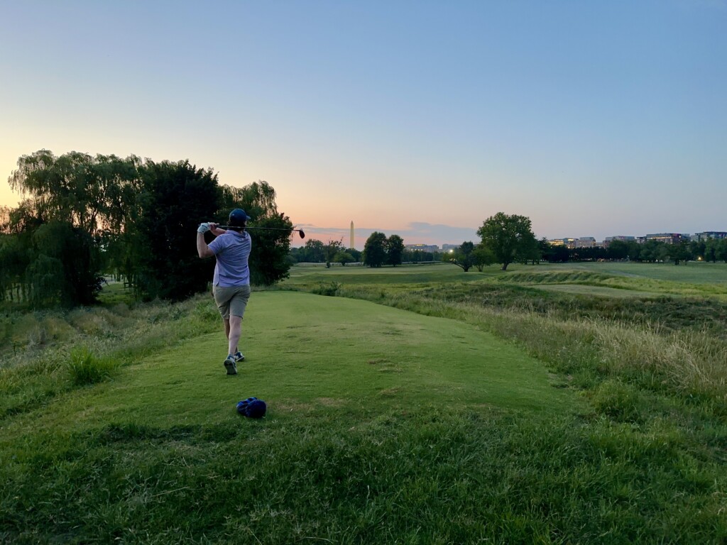 Golfer following through on a tee shot from the elevated 18th tee at East Potomac Golf Links Blue Course, with the Washington Monument and DC skyline on the horizon at dusk.