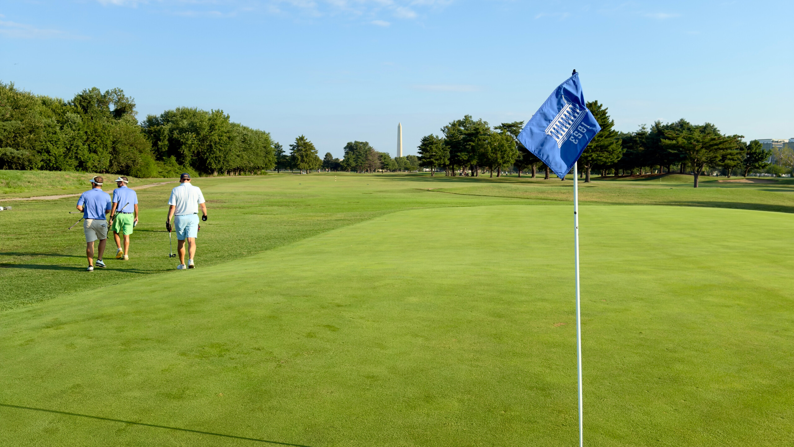 Looking back down the 2nd fairway at East Potomac Golf Links Blue Course from behind the green, with three golfers walking off, a blue flagstick in the foreground, and the Washington Monument on the horizon.