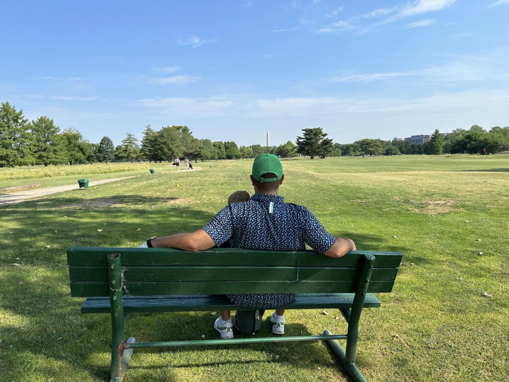 A golfer sits on a weathered green bench near the 8th tee at East Potomac Golf Links - Blue Course, looking out across the property toward the Washington Monument on the horizon.
