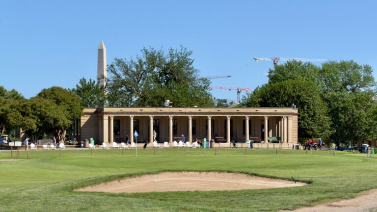 The federal-style colonnaded clubhouse at East Potomac Golf Links viewed from the right rough on the 9th hole, with the Washington Monument visible behind it and a bunker in the foreground.