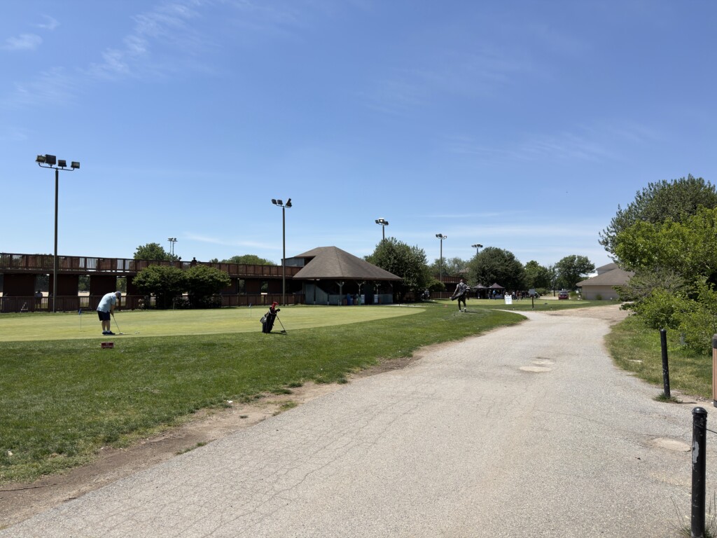 Two-tier driving range at East Potomac Golf Links with golfers practicing on the putting greens out front under a clear June sky