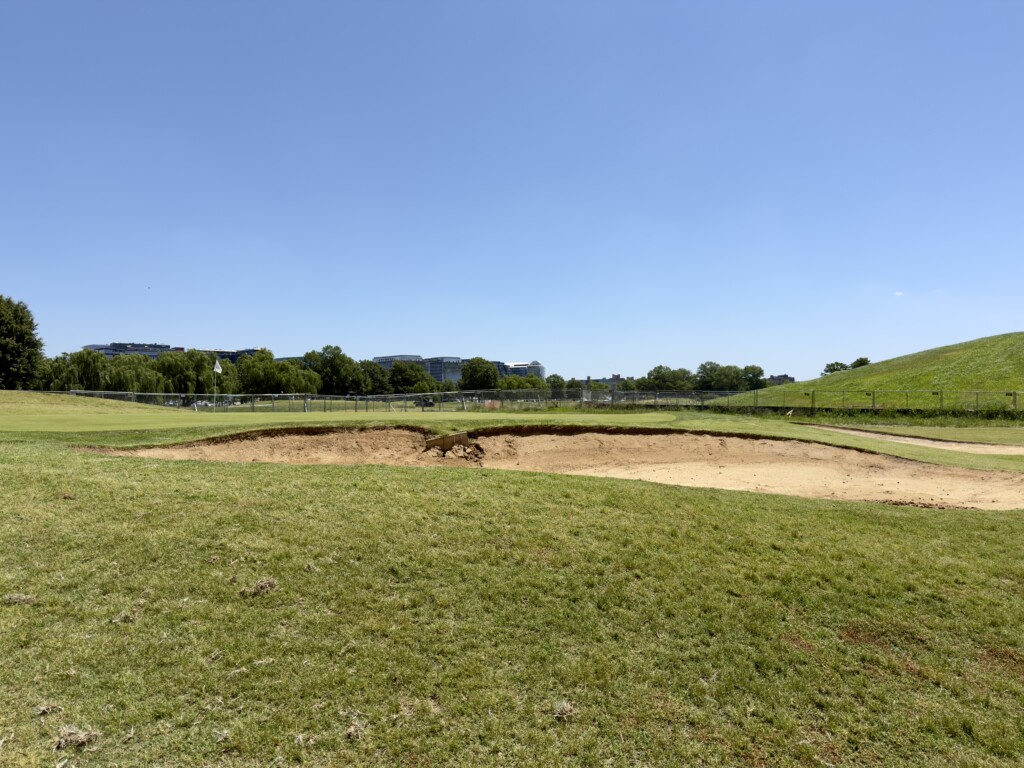 Torn-up greenside bunker at the 5th green of East Potomac White Course, grassy mound and DC skyline beyond