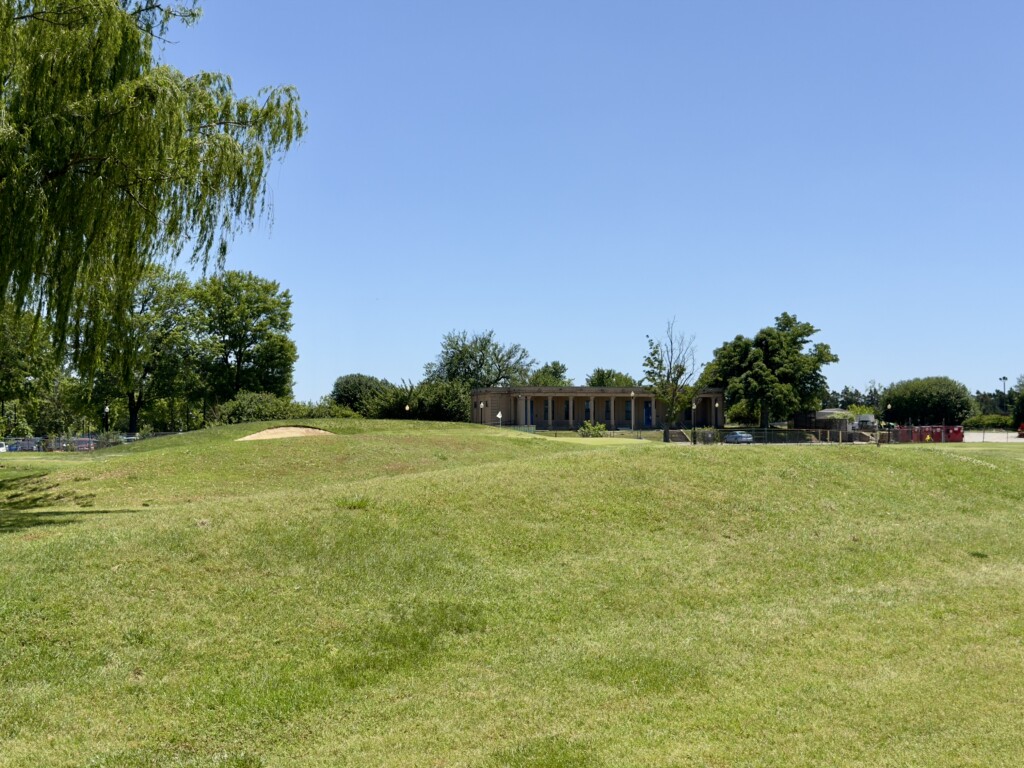 Grassy mounding on the left rough of the 7th at East Potomac White Course, bunker dug into the slope, colonnade building beyond